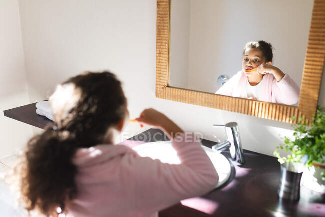 Mixed race girl looking at mirror and brushing teeth in bathroom. domestic lifestyle and spending quality time at home. — Stock Photo