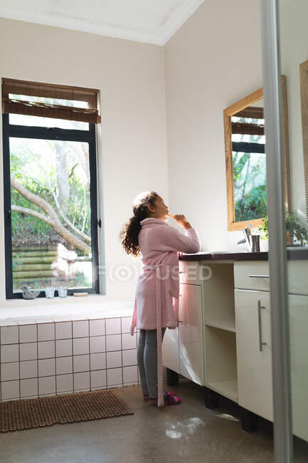 Chica de raza mixta mirando al espejo y cepillándose los dientes en el baño. estilo de vida doméstico y pasar tiempo de calidad en casa. - foto de stock