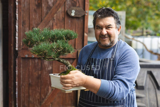 Ritratto di giardiniere maschio caucasico sorridente che tiene un albero di bonsai al centro del giardino. specialista che lavora presso vivaio bonsai, attività orticola indipendente. — Foto stock