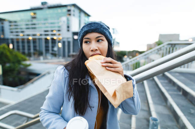 Asiatin isst und hält Kaffee zum Mitnehmen auf der Straße. Unabhängige junge Frau in der Stadt unterwegs. — Stockfoto