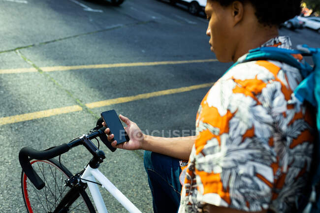 Hombre afroamericano en la ciudad sentado en bicicleta y usando un teléfono inteligente. nómada digital sobre la marcha, fuera y alrededor de la ciudad. - foto de stock