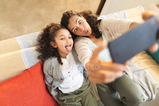 Mixed race mother and daughter sitting on sofa doing funny faces, taking selfie. domestic lifestyle and spending quality time at home. — Stock Photo