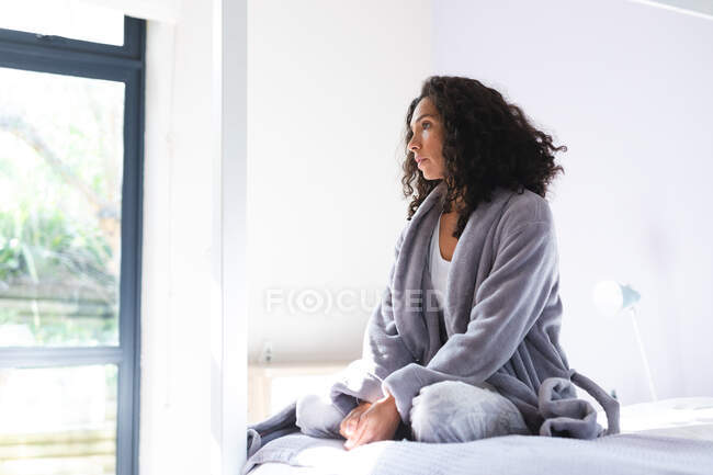 Happy thoughtful mixed race woman sitting on bed and looking through window. domestic lifestyle and spending quality time at home. — Stock Photo