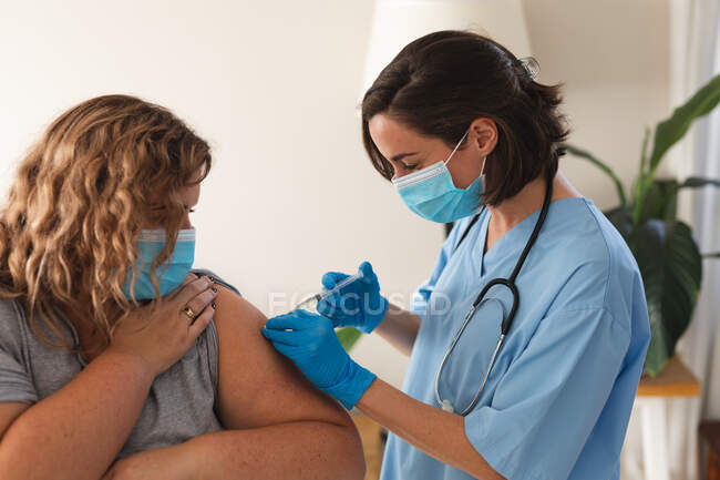 Caucasian female doctor wearing face mask vaccinating female patient at home. medical and healthcare services home visiting during coronavirus covid 19 pandemic. — Stock Photo
