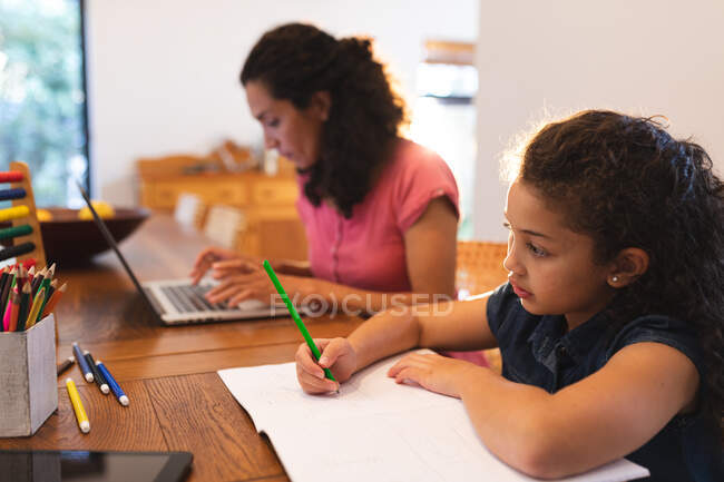 Madre e figlia di razza mista sedute a tavola, che lavorano su laptop e disegno. stile di vita domestico e trascorrere del tempo di qualità a casa. — Foto stock