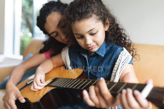 Raza mixta madre e hija sentadas en el sofá y tocando la guitarra. estilo de vida doméstico y pasar tiempo de calidad en casa. - foto de stock