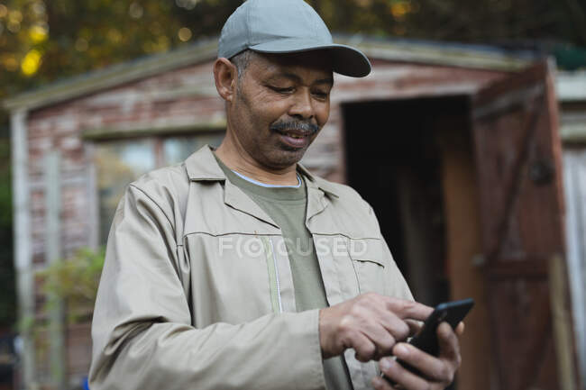 Jardinero masculino afroamericano usando smartphone en el centro del jardín. especialista en vivero de plantas bonsái, empresa hortícola independiente. - foto de stock