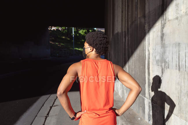 Ajuste hombre afroamericano haciendo ejercicio en la ciudad con máscara facial. aptitud física y estilo de vida urbano activo al aire libre durante coronavirus covid 19 pandemia. - foto de stock