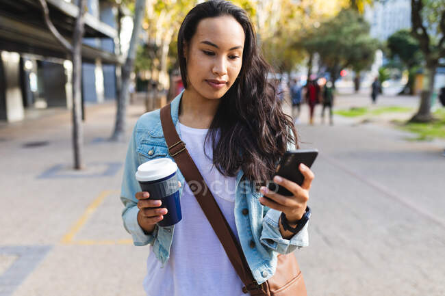 Donna asiatica utilizzando smartphone e tenendo il caffè da asporto per strada. indipendente giovane donna in giro per la città. — Foto stock
