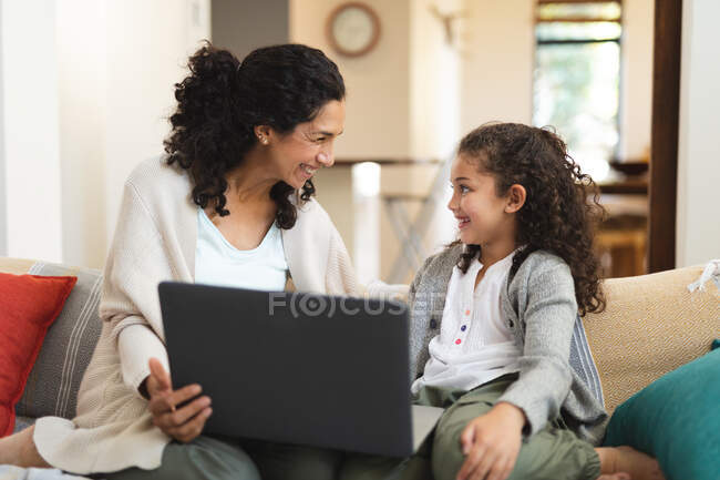 Sonriente madre de raza mixta y su hija sentados en el sofá y el uso de ordenador portátil. estilo de vida doméstico y pasar tiempo de calidad en casa. - foto de stock