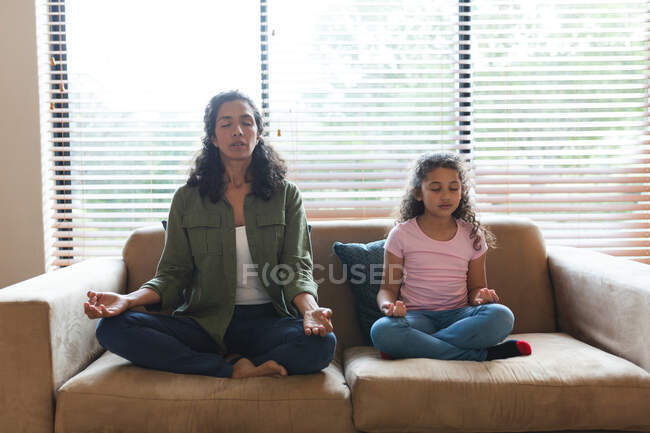 Mixed race mother and daughter sitting on sofa and meditating. domestic lifestyle and spending quality time at home. — Stock Photo