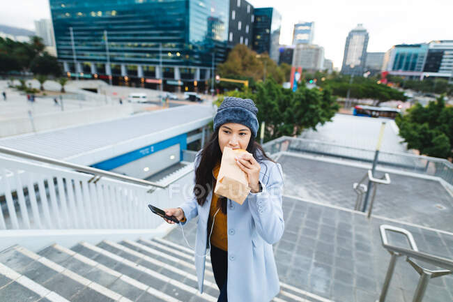 Mulher asiática comendo e segurando smartphone na rua. jovem independente para fora e sobre na cidade. — Fotografia de Stock