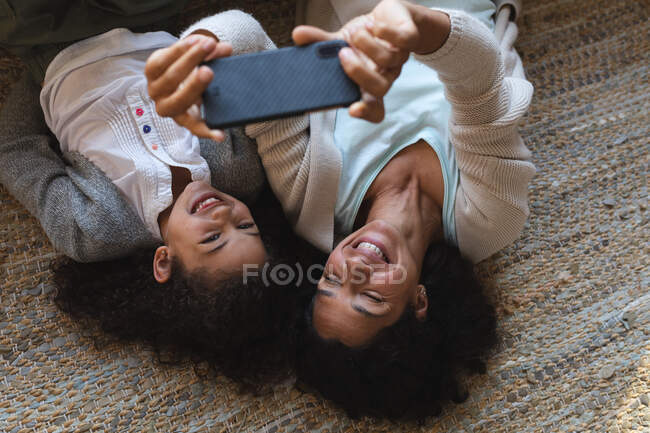Misto razza madre e figlia sdraiati su un tappeto, prendendo selfie. stile di vita domestico e trascorrere del tempo di qualità a casa. — Foto stock