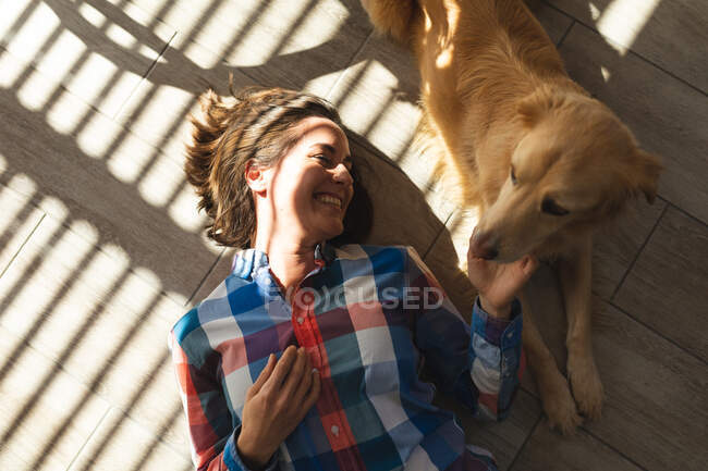 Белая женщина, лежащая на полу, улыбается и гладит собаку. бытовой образ жизни, свободное время дома. — стоковое фото