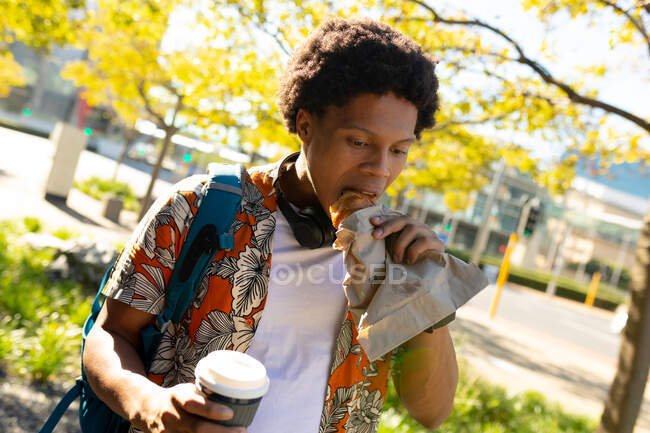 Homem afro-americano na cidade a comer e a segurar café. nômade digital em movimento, para fora e sobre na cidade. — Fotografia de Stock
