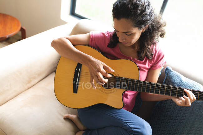 Gemischte Rassenfrau sitzt auf Sofa und spielt Gitarre. Lebensstil und hochwertige Zeit zu Hause verbringen. — Stockfoto