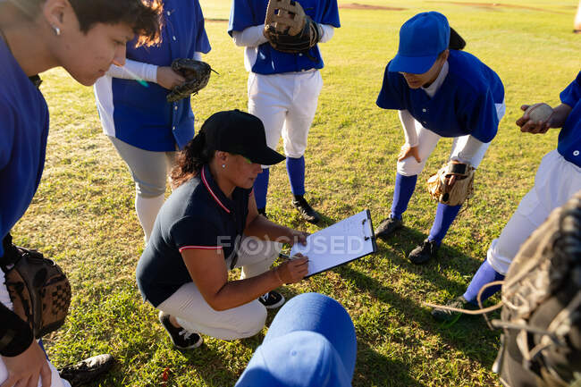 Разнообразная группа женщин-бейсболистов в обнимку вокруг приседающих женщин-тренеров с планшетом. женская бейсбольная команда, спортивная подготовка и тактика игры. — стоковое фото