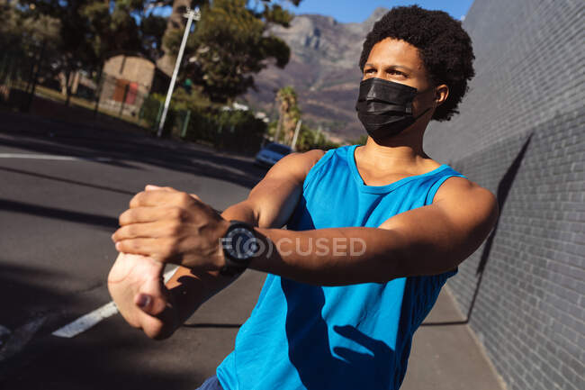 Ajuste hombre afroamericano haciendo ejercicio en la ciudad usando mascarilla, estirándose en la calle. aptitud física y estilo de vida urbano activo al aire libre durante coronavirus covid 19 pandemia. - foto de stock
