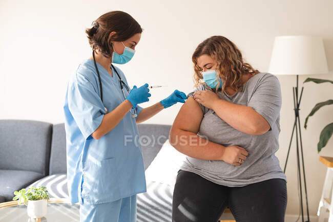 Doctora caucásica que usa mascarilla facial vacunando a una paciente femenina en casa. servicios médicos y sanitarios visita domiciliaria durante la pandemia de coronavirus covid 19. - foto de stock