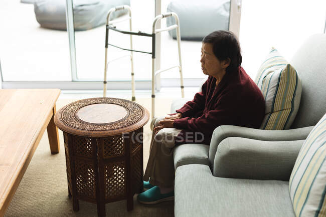 Triste mujer asiática mayor en casa sentada en el sofá en la sala de estar. estilo de vida senior, pasar tiempo en casa. - foto de stock