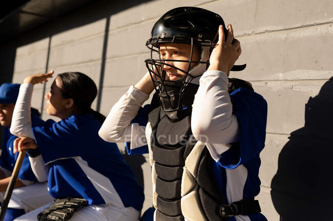 Белая женщина-бейсболистка сидит на скамейке, а товарищи по команде надевают шлем перед игрой. женская бейсбольная команда, подготовленная и ожидающая игры. — стоковое фото