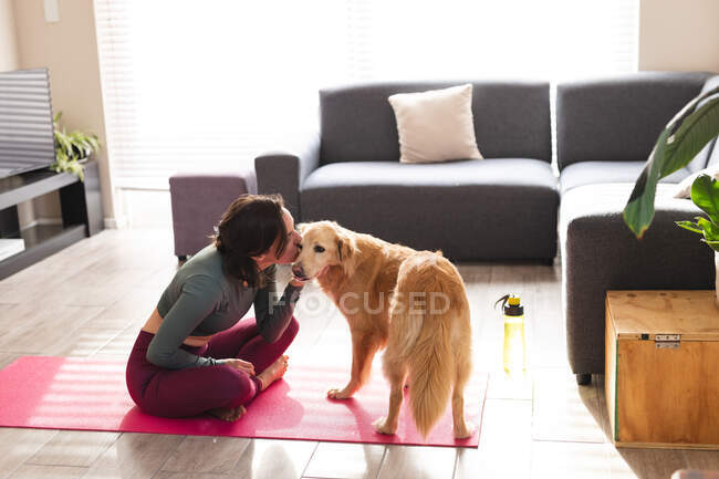 Mujer caucásica practicando yoga, sentada en una esterilla de yoga besando a su perro. estilo de vida doméstico, pasar tiempo libre en casa. - foto de stock