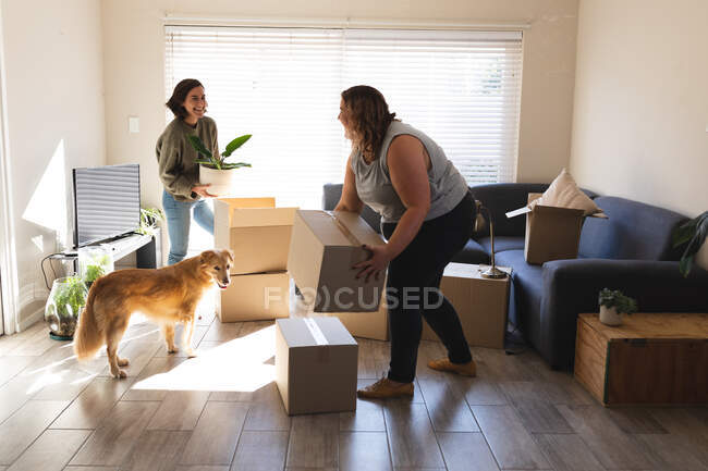 Lesbian couple with dog smiling and holding boxes during moving house. domestic lifestyle, spending free time at home. — Stock Photo