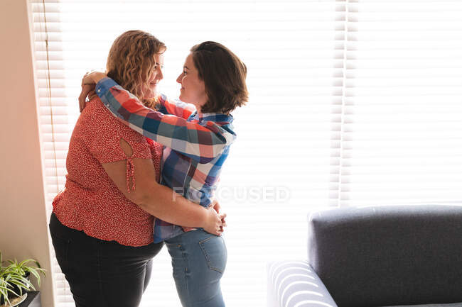 Happy lesbian couple embracing and smiling next to window. domestic lifestyle, spending free time at home. — Stock Photo