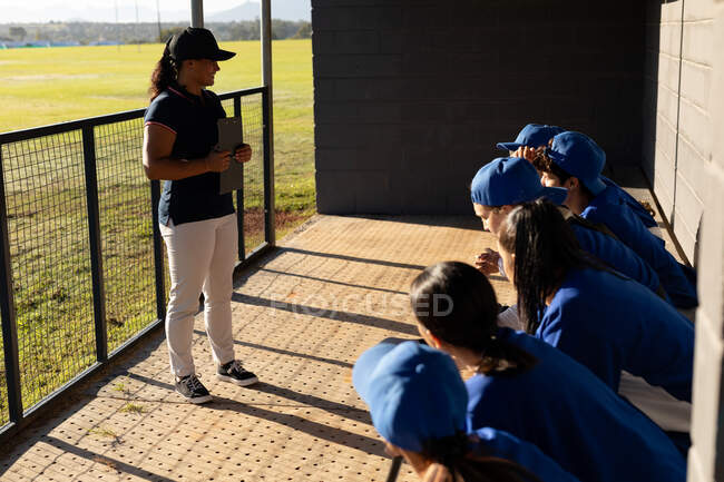 Разнообразная группа женщин-бейсболистов, сидящих на скамейке запасных, слушающих женщин-тренеров перед игрой. женская бейсбольная команда, спортивная подготовка и тактика игры. — стоковое фото