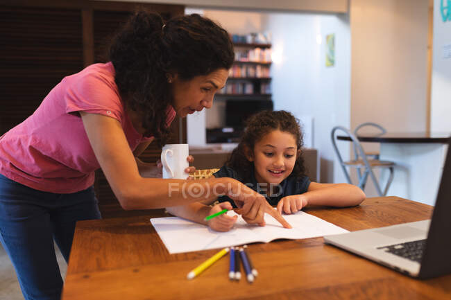 Mutter mit gemischter Rasse hält Kaffee und hilft ihrer Tochter bei den Hausaufgaben. Lebensstil und hochwertige Zeit zu Hause verbringen. — Stockfoto