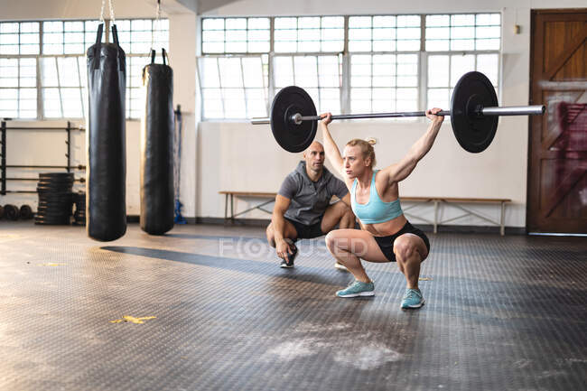 Entrenador masculino caucásico instruyendo a la mujer haciendo ejercicio en el gimnasio, levantando pesas. entrenamiento cruzado de fuerza y fitness para boxeo. - foto de stock