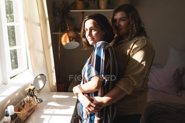 Coppia lesbica abbracciando sorridente e guardando attraverso la finestra. stile di vita domestico, trascorrere il tempo libero a casa. — Foto stock