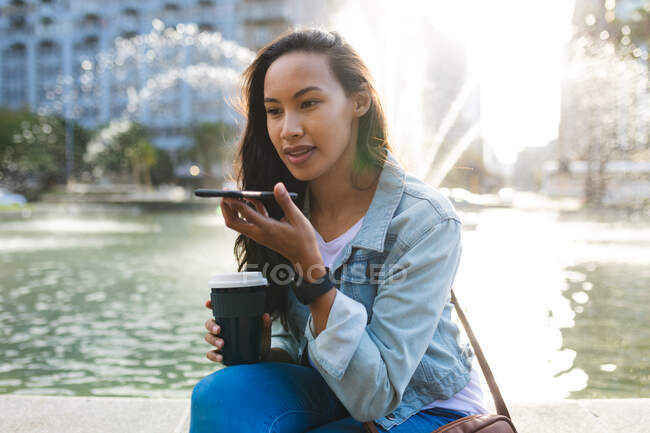 Asiatin im Smartphone-Gespräch und Kaffee zum Mitnehmen im sonnigen Park. Unabhängige junge Frau in der Stadt unterwegs. — Stockfoto