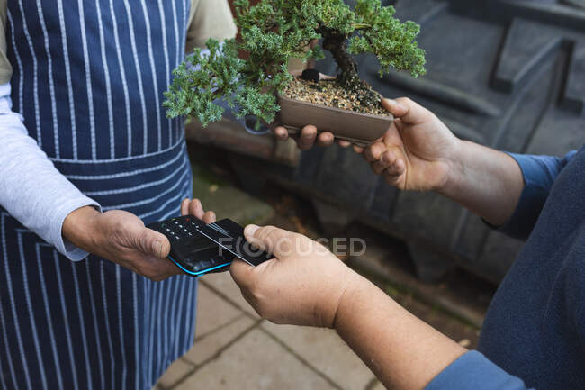 Sezione centrale di due uomini diversi che utilizzano pagamenti senza contatto al centro del giardino. specialisti che lavorano presso vivaio bonsai, attività orticola indipendente. — Foto stock