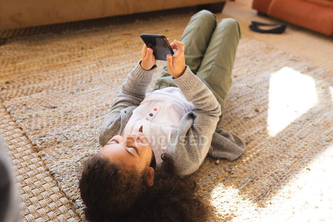 Дівчина змішаної раси лежить на килимі, використовуючи смартфон. домашній спосіб життя і якісний час вдома . — стокове фото