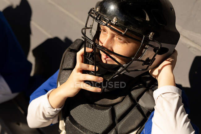 Jugadora de béisbol caucásica sentada en el banco poniéndose el casco del receptor antes del partido. equipo femenino de béisbol, preparado y esperando el juego. - foto de stock