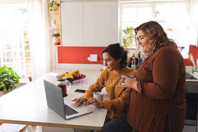 Лесбиянки улыбаются и используют ноутбук на кухне. бытовой образ жизни, свободное время дома. — стоковое фото