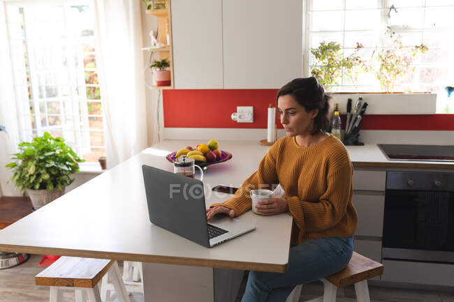 Mujer caucásica beber café y el uso de ordenador portátil en la cocina. estilo de vida doméstico, pasar tiempo libre en casa. - foto de stock