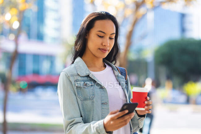 Donna asiatica utilizzando smartphone e tenendo il caffè da asporto nel parco soleggiato. indipendente giovane donna in giro per la città. — Foto stock