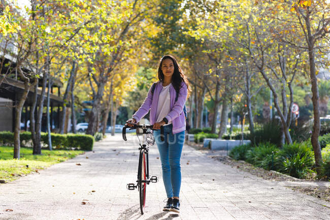 Ritratto di donna asiatica sorridente che ruota in bicicletta e guarda la macchina fotografica nel parco soleggiato. indipendente giovane donna in giro per la città. — Foto stock