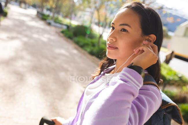Улыбающаяся азиатка катается на велосипеде и пользуется наушниками в солнечном парке. независимая молодая женщина в городе. Идентификация — стоковое фото