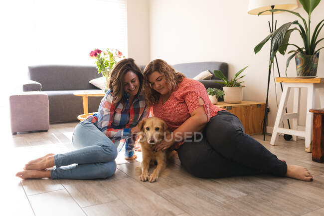 Feliz pareja lesbiana sentada en el suelo abrazando y sonriendo con su perro. estilo de vida doméstico, pasar tiempo libre en casa. - foto de stock