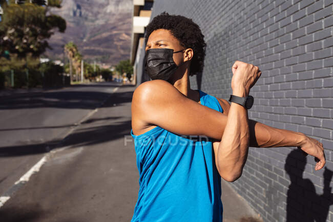Fit african american man exercising in city wearing face mask, stretching in the street. fitness and active urban outdoor lifestyle during coronavirus covid 19 pandemic. — Stock Photo