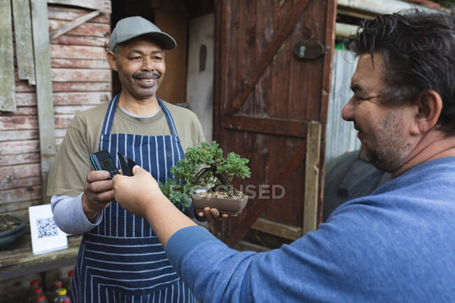 Два різноманітних чоловіки використовують безконтактні платежі в садовому центрі. спеціалісти, що працюють в розпліднику бонсайських рослин, незалежний садівничий бізнес . — стокове фото