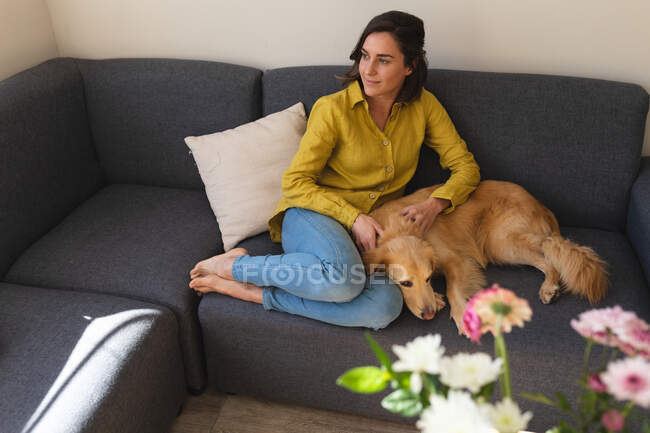 Femme blanche portant une chemise jaune et assise sur le canapé avec chien. mode de vie domestique, passer du temps libre à la maison. — Photo de stock