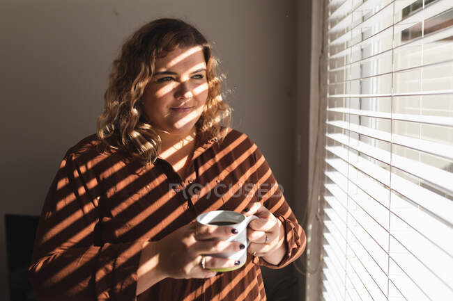 Donna caucasica sorride e beve caffè vicino alla finestra. stile di vita domestico, trascorrere il tempo libero a casa. — Foto stock