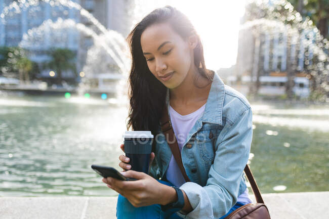 Mujer asiática usando smartphone y sosteniendo café para llevar en el soleado parque. mujer joven independiente fuera y alrededor de la ciudad. - foto de stock