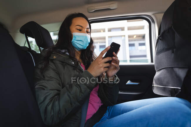 Asiatin mit Gesichtsmaske sitzt im Taxi und benutzt Smartphone. Unabhängige junge Frau während der Coronavirus-Pandemie 19 in der Stadt unterwegs. — Stockfoto