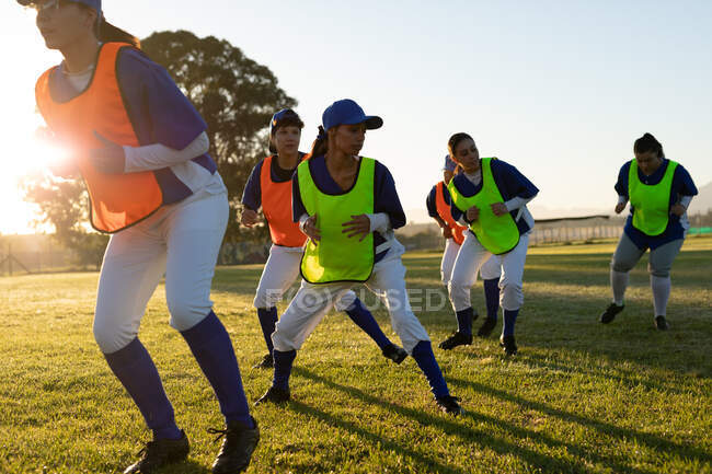 Groupe diversifié de joueuses de baseball portant des dossards colorés s'entraînant sur le terrain au lever du soleil. équipe féminine de baseball, entraînement sportif et tactiques de jeu. — Photo de stock