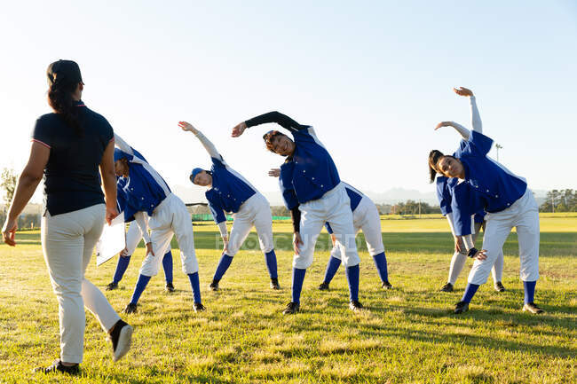 Разнообразная группа женщин-бейсболистов с тренером, разогревающаяся на поле, растянувшаяся от пояса. женская бейсбольная команда, спортивные тренировки, сплоченность и приверженность. — стоковое фото